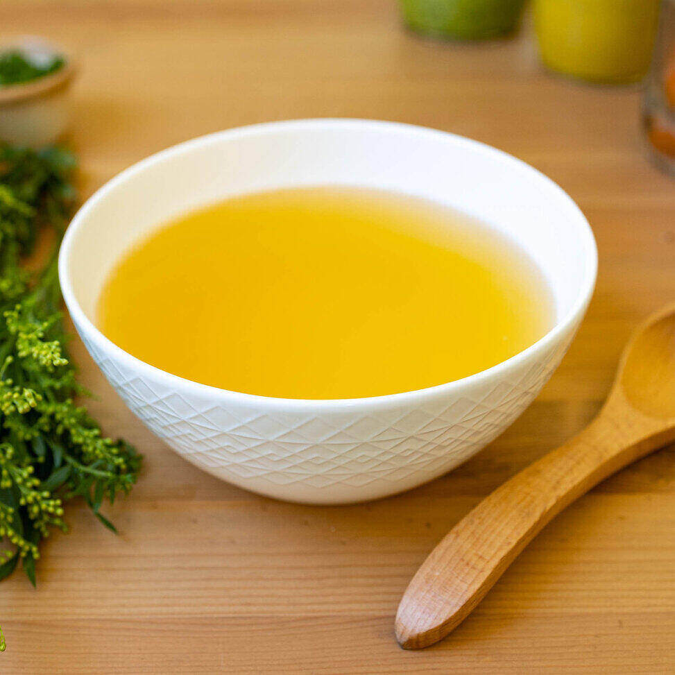 A white ceramic bowl with a geometric design sits in the middle of the photo on a wood table with a wooden spoon on its right and a bunch of herbs on its left. The soup is bone broth - so a clear brown liquid.