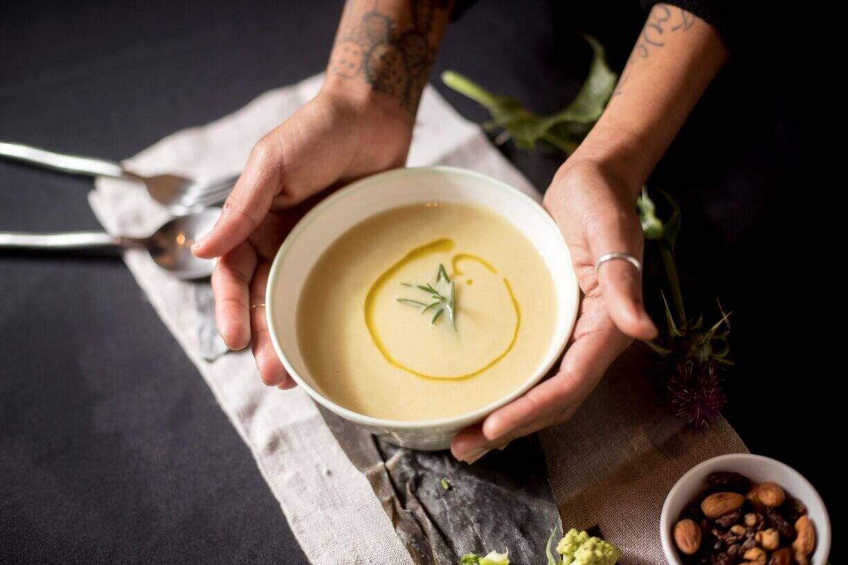 Two hands cup a bowl of nourishing soup - soup is light yellow in color with a drizzle of oil on it and a few herbs floating in the middle. The bowl of soup sits on a beige linen placemat with a bowl of nuts to the right and silverware to the left. 