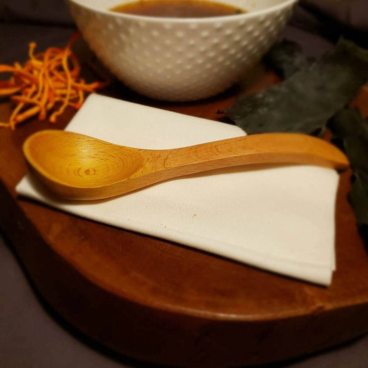 Purpose and Hope suggests using a wooden spoon when eating soups in order to avoid a metallic taste, especially post medical treatment. Pictured here is a wooden soup spoon on a white linen napkin sitting on a wood board with a bowl of soup in the background. 