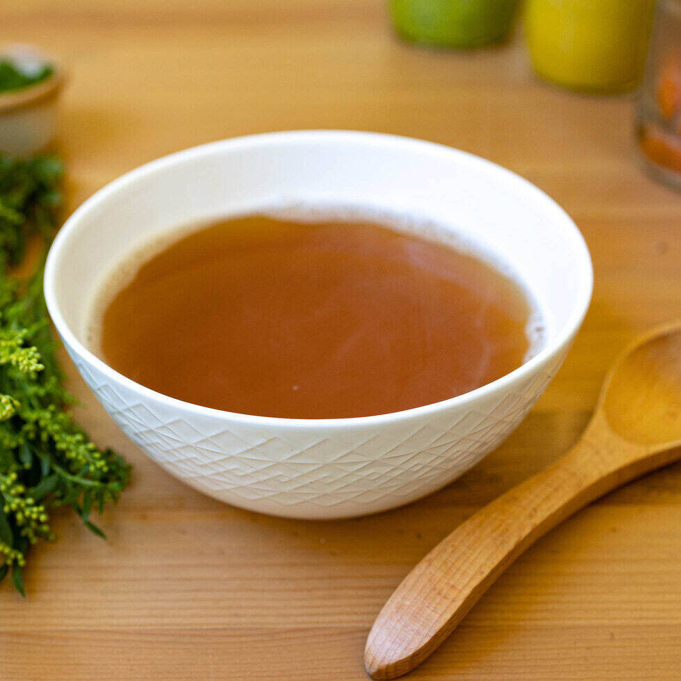 A white ceramic bowl with a geometric design sits in the middle of the photo on a wood table with a wooden spoon on its right and a bunch of herbs on its left. The soup is bone broth - so a clear brown liquid.
