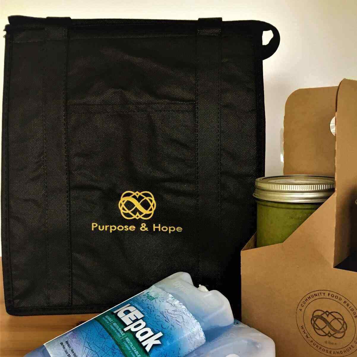 A black tote bag with the Purpose and Hope logo on it sits behind two ice packs and a cardboard drink holder holding a jar of green soup. 