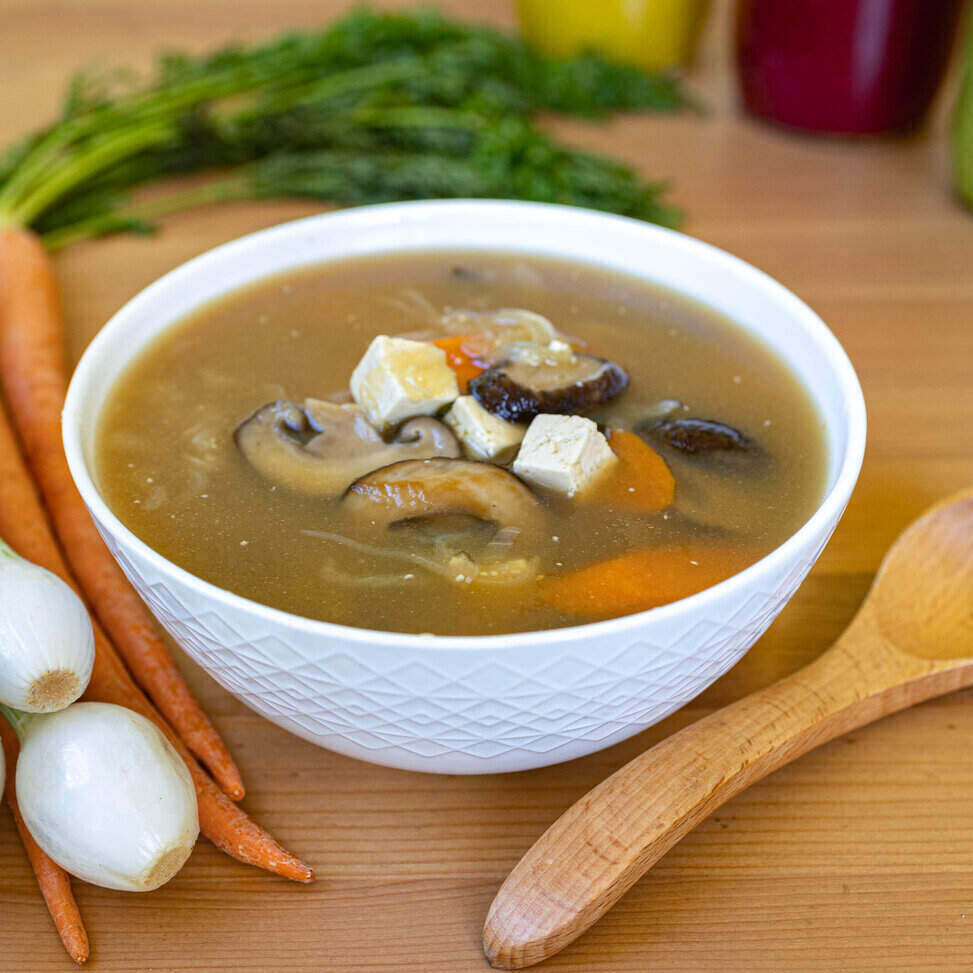 A white ceramic bowl with a geometric design sits in the middle of the photo on a wood table with a wooden spoon on its right and a bunch of herbs on its left. The soup is bone broth - so a clear brown liquid with mushrooms, tofu, and root vegetables. 