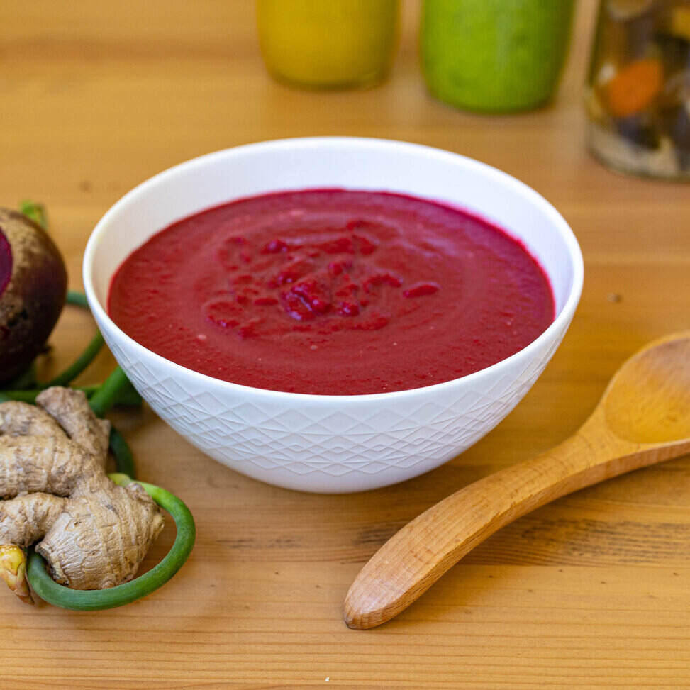 A white ceramic bowl with a geometric design sits in the middle of the photo on a wood table with a wooden spoon on its right and a bunch of herbs on its left. The soup is a blended soup of red beet, coconut milk and ginger so it is a bright red color. 