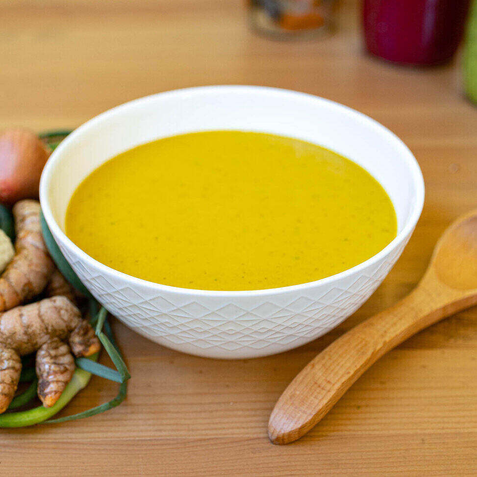 A white ceramic bowl with a geometric design sits in the middle of the photo on a wood table with a wooden spoon on its right and a bunch of herbs on its left. The soup is a blended soup of cauliflower and turmeric so it is yellow in color. 