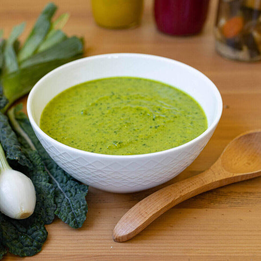 A white ceramic bowl with a geometric design sits in the middle of the photo on a wood table with a wooden spoon on its right and a bunch of herbs on its left. The soup is a blended soup of broccoli and greens so it is green in color. 