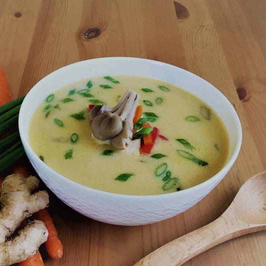 A white ceramic bowl with a geometric design sits in the middle of the photo on a wood table with a wooden spoon on its right and a bunch of herbs on its left. The soup is a blended soup that is yellow in color with vegetables and herbs floating in it. 