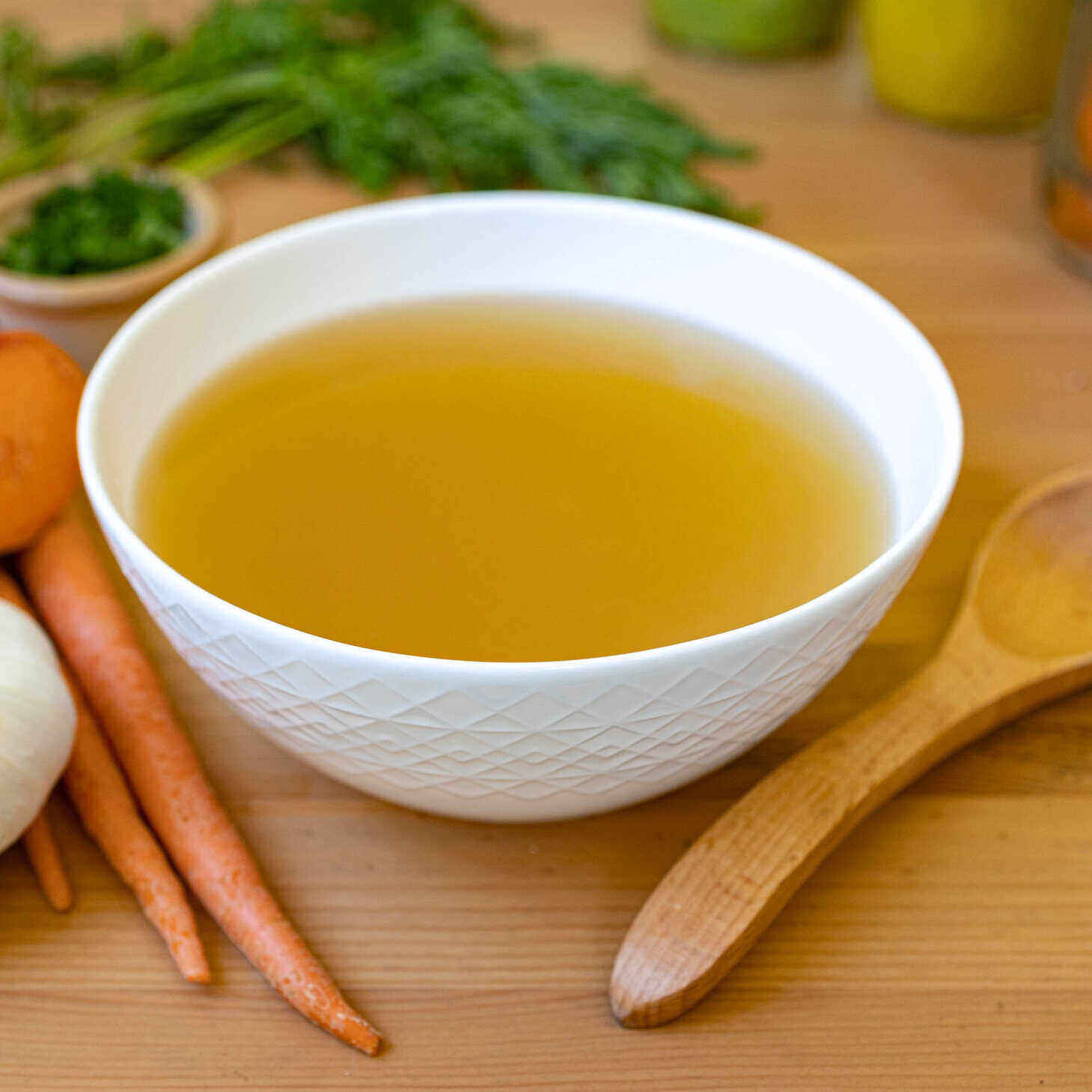 A white ceramic bowl with a geometric design sits in the middle of the photo on a wood table with a wooden spoon on its right and a bunch of herbs on its left. The soup is vegetable broth - so a clear brown liquid.